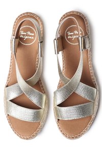 Espadrilles métalliques dorées avec des lanières croisées, semelle plate et bord en jute tissé. La semelle intérieure est en cuir marron avec un logo marqué.