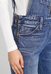 Denim overalls in light blue with a textured finish. Features a front zipper and pockets, complemented by orange stitching details.