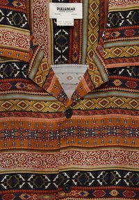 Kortærmet skjorte med et farverigt geometrisk mønster i nuancer af orange, brun og sort, med nedfoldet krave og knaplukning.