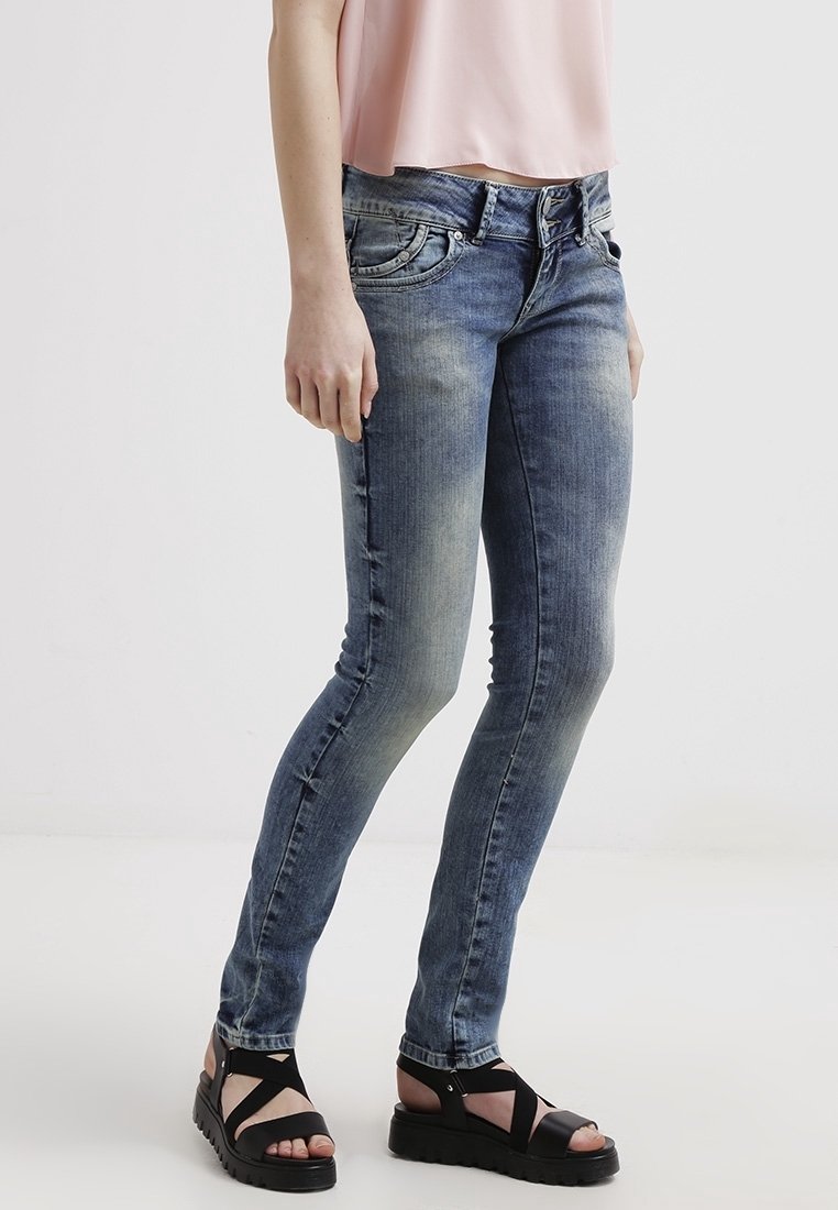 Woman wearing faded skinny blue jeans, a pink sleeveless top, and black platform sandals standing against a plain white background.