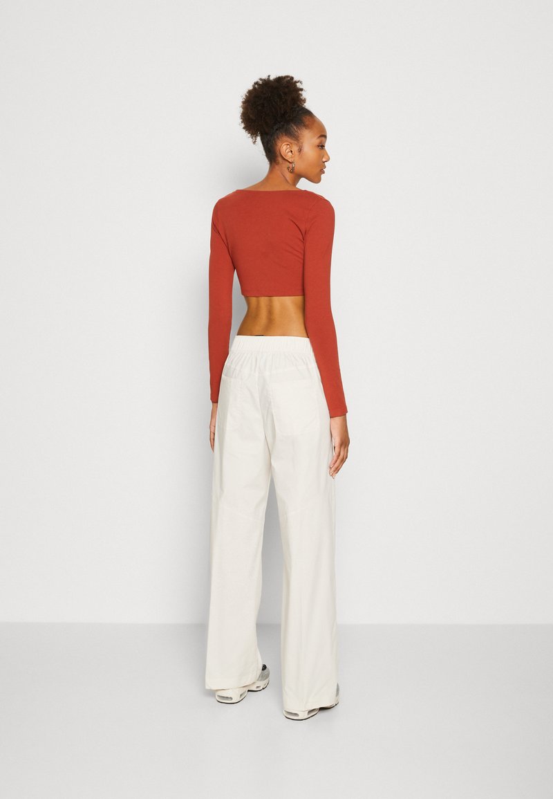Rostfarbenes langärmeliges Crop-Top, kombiniert mit weißen, hoch taillierten, weiten Hosen. Das Top hat einen quadratischen Ausschnitt und eine figurbetonte Passform, während die Hosen eine lässige Passform aufweisen.