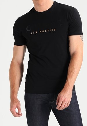 Homme portant un t-shirt noir avec l'inscription "Los Angeles" et un jean bleu foncé, debout devant un fond uni clair.