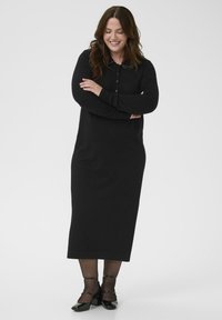 Robe longue noire à manches longues avec patte de boutonnage, coupe décontractée, tissu texturé et silhouette droite. Associée à des chaussures noires à talons épais.