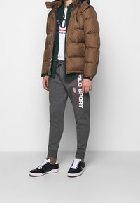 Hnědá puffová bunda, šedé tepláky s potiskem "POLO SPORT", bílý tričko, černé tenisky s bílými šňůrkami, kontrastní textury a barvy.