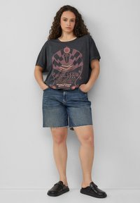 T-shirt oversized gris foncé avec impression graphique en rouge passé, associé à un short en denim bleu arrivant aux genoux avec un ourlet effiloché et des chaussures noires à enfiler.
