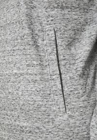 Šedá tkanina s texturou a melírovaným vzorem; obsahuje boční kapsu, která se integruje do materiálu, což nabízí uvolněný a funkční design.