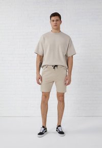 T-shirt bege de algodão mangas curtas com bolso no peito, combinada com calções bege a condizer e ténis pretos. Design simples, corte descontraído.