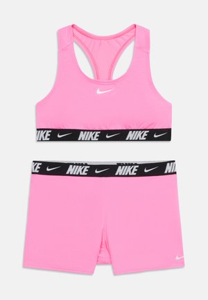 Rozā Nike sporta krūšturis un atbilstoši šorti ar melnām elastīgām lentēm, uz kurām ir balti Nike logotipi un spēles zīmes.