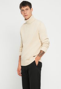 Cremefarvet turtleneck sweater lavet af tekstureret strikstof, med ribbet kant og manchetter, parret med sorte bukser. Diskret mærke detail på siden.
