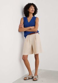 Haut sans manches en tricot bleu avec un col en V, shorts plissés beiges et sandales gris clair. Complété par une ceinture tressée marron, sur un fond uni.