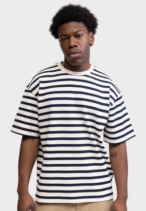 Giovane uomo di colore che indossa una maglietta a righe orizzontali bianche e blu navy a maniche corte, in piedi davanti a uno sfondo chiaro uniforme.