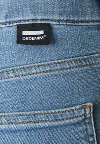 Blå denimtyg med gula sömmar, som visar en ficka och en svart etikett med texten "DRDENIM" och ett vitt horisontellt streck ovanför texten.