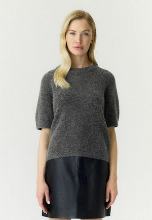 Donna con i capelli biondi e lunghi che indossa un maglione grigio a maniche corte e una gonna di pelle nera, in piedi davanti a uno sfondo semplice.