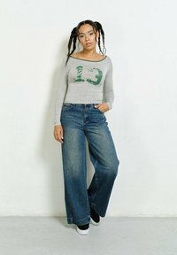 Gray long-sleeve top with green printed "19" and contrasting neckline, paired with wide-leg blue jeans and black shoes with white soles.