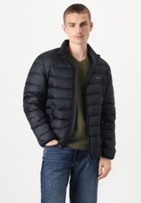 Giacca a vento nera con design trapuntato, chiusura con zip e tasche laterali, abbinata a una maglietta V-neck verde oliva e jeans blu.