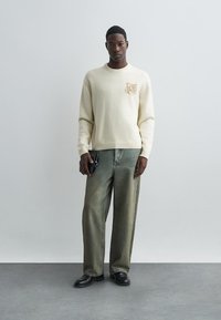 Kermato paita, jossa on uritettut hihansuut ja tekstuurinen vaaleanruskea läntti. Yhdistettynä vaaleanvihreisiin väljiksi farkkuihin ja mustiin kenkiin, pitäen pientä mustaa lompakkoa.