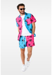 Kortærmet skjorte med knapper i blå og pink med sort palmeprint, parret med matchende badebukser, båret med en hvid T-shirt og solbriller.