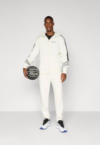 Traje de chándal color crema con acentos en negro y gris, sudadera con cremallera y logo, y pantalones ajustados. Balón de baloncesto negro en la mano. Zapatillas blancas.