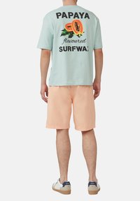 Miętowa koszulka z napisem "PAPAYA flavoured SURFWAX" i grafiką na plecach, w połączeniu z brzoskwiniowymi szortami i białymi trampkami.