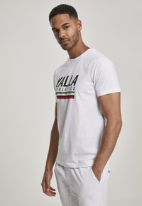 Bílý bavlněný tričko s nápisem "YALLA ATHLETICS" vytištěným tučným černým písmem, doplněné o zelené a červené vodorovné pruhy na spodním lemu.