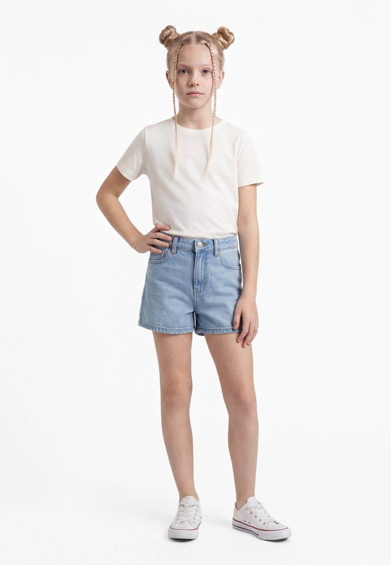 Heledate juustega tüdruk, kellel on topelttutinöörid ja punutud kiud, kannab valget t-särki, siniseid teksapükse ja valgeid tossusid ning seisab ühe käega puusal.