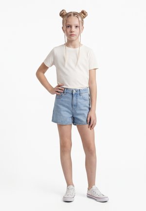 Blondinka z dvojnimi figami in pletenimi prameni nosi belo majico, modre jeans kratke hlače in bele superge, stoji z eno roko na boku.