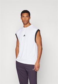 Weißes ärmelloses Sportshirt mit schwarzen Akzenten, mit einem Logo auf der Brust. Kombiniert mit dunklen Hosen. Einfaches Design und lockere Passform.