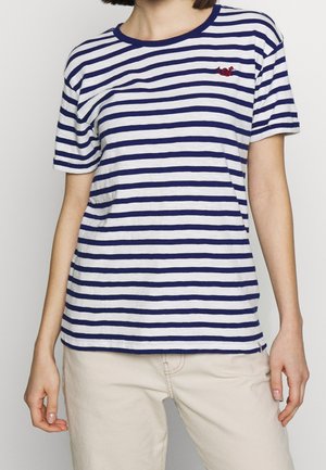 Vrouw draagt een wit met marineblauw gestreept T-shirt met korte mouwen en een klein rood geborduurd detail op de borst, met lichtbeige broek.