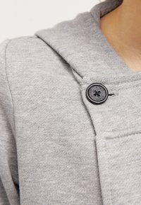Sweatshirt gris avec une texture douce, présentant une grande capuche et un détail de bouton noir à l'avant. Le tissu semble léger et décontracté.