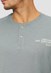 Svijetlo siva pamučna henley majica s dva dugmeta na izrezanju i malim bijelim tekstom na lijevoj strani prsa. Mekana textura s suptilnim uzorkom.