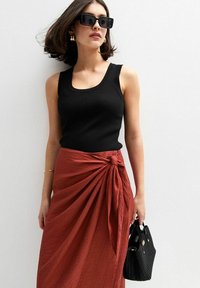 Top a coste nero abbinato a una gonna avvolgente testurizzata color ruggine con dettaglio a nodo. Gli accessori includono occhiali da sole neri e una borsa nera piccola.