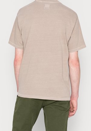 Beiges Baumwoll-T-Shirt mit kurzen Ärmeln, rundem Ausschnitt und sichtbaren Nähten. Wird mit grüner Hose getragen und zeigt eine legere Passform.