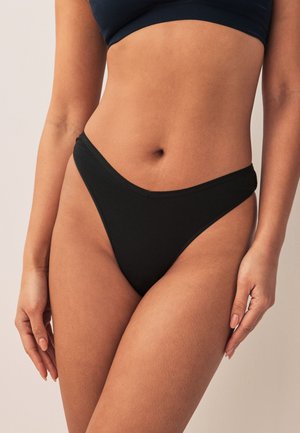 Nainen, joka käyttää mustaa korkeavyötäröistä alusvaatetta ja tummaa lyhyttä toppia neutraalia taustaa vasten, näyttäen vyötärön ja lantion.