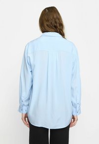 Camicia a maniche lunghe azzurro chiaro con colletto, polsini con bottoni e dettagli con pieghe sul retro. Texture liscia con vestibilità rilassata e orlo curvo.
