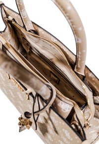 Bolso de mano beige con superficie texturizada, herrajes dorados, cierre de cremallera y múltiples compartimentos. Presenta un interior con un patrón y un charm decorativo.