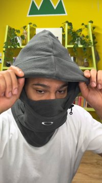 Glugă gri din fleece cu o caracteristică de acoperire a feței și șnur reglabil. Include un logo pe partea din față. Purtat peste un tricou alb.