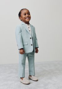 Lichtblauw pak van zachte stof, met een colbert in een enkele rij met twee knopen, gecombineerd met een beige overhemd met kraag en grijze mocassins.