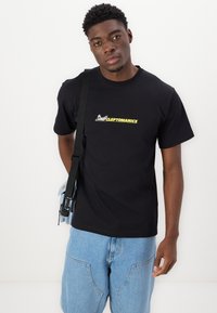 Black cotton t-shirt featuring a front graphic in yellow and white. Model wears light blue denim shorts and holds a blue bag with a strap.