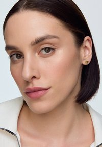 Złote kolczyki w kształcie serca na osobie z średnio brązowymi włosami, gładką skórą, naturalnym makijażem i noszącą jasny top.