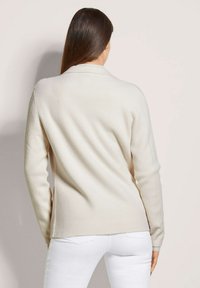 Strickcardigan in Cremefarbe mit weicher Textur, ausgestattet mit langen Ärmeln, einem Kragen und seitlichen Schlitzen, getragen über weißen Jeans.