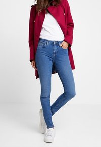 Jeans ajustados en denim azul medio con una textura suave, combinados con una camiseta blanca y un abrigo largo burdeos. Zapatillas blancas completan el look.