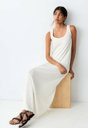 Femme aux cheveux foncés dans une longue robe blanche sans manches assise sur un bloc de bois, portant des sandales noires à clous, regardant sur le côté.