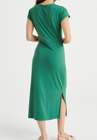 Zelené šaty ke kolenům vyrobené z měkké tkaniny. Mají krátké rukávy a boční rozparek. Hladká textura s dobře padnoucím pasem a uvolněným střihem.
