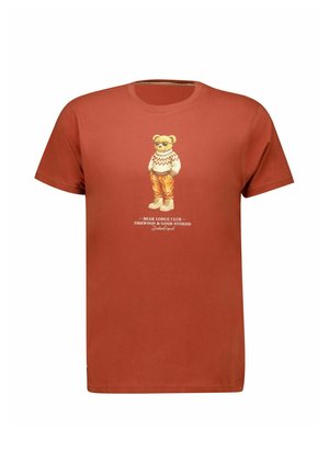 T-shirt à manches courtes couleur rouille avec un dessin d’un ours portant un pull, un pantalon et des bottes, debout les mains dans les poches.