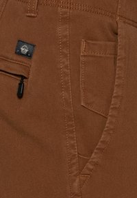 Pantalon en tissu marron avec une poche avant, une petite poche zippée et des coutures contrastées. Le matériau a une texture lisse avec un design durable.