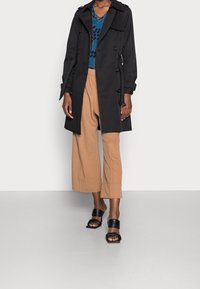 Schwarzer Trenchcoat mit doppelt brechenden Knöpfen, getragen über einem blauen, gemusterten Oberteil und beigen, weit geschnittenen Hosen, kombiniert mit schwarzen Rutsch-Sandalen.