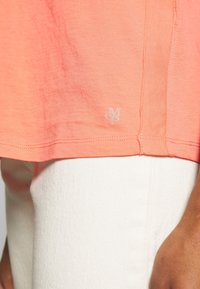 Korálové tričko s krátkým rukávem, které má decentní logo na lemu, hladkou texturu a rovný střih. V kombinaci s bílými kalhotami.