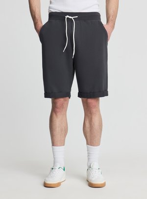 Mężczyzna ubrany w czarne szorty ze ściągaczem, białe skarpetki sportowe i białe sneakersy z zielonymi akcentami, stojący z rękami w kieszeniach.