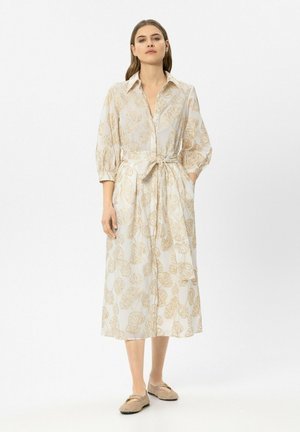 Frau trägt ein beiges Midikleid mit Blättermuster, 3/4-Ärmeln, Taillengürtel und gewebte beige Ballerinas, steht vor einem weißen Hintergrund.