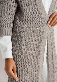 Hellgraue gestrickte Strickjacke mit offenem Design, strukturierten Mustern und gerippten Kanten, darunter ein langärmliges weißes Hemd.